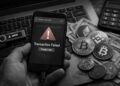 Smartphone displaying a ‘transaction failed, and funds lost' warning, beside physical cryptocurrency coins and dollar bills on a laptop.