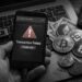 Smartphone displaying a ‘transaction failed, and funds lost' warning, beside physical cryptocurrency coins and dollar bills on a laptop.